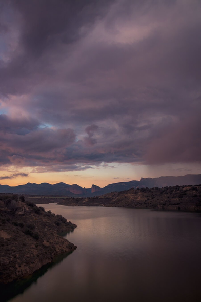 Vertical photograph showing a landscape at the end of the sunset in violet and orange tones. In the foreground a large reservoir. In the background a line of mountains. And above them, occupying more than half of the photograph, a sky with large storm clouds.