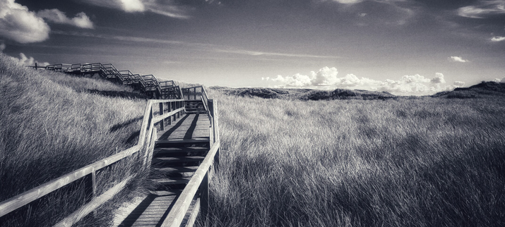 Schwarz weiß Bild. Abgebildet ist ein Holzsteg welcher sich durch Dünen schlängelt. Der Steg führt in Richtung links bergauf und ist durch mehrere Treppenläufe durchtrennt. Ein paar Wolken suchen sich ihren Weg am Himmel. 