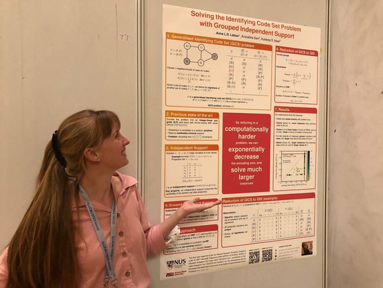 Me, a young woman with long blonde hair wearing a pink blouse, standing in front of an academic poster, smilingly posing as if I am explaining it to someone. The poster is titled "Solving the Identifying Code Set Problem with Grouped Independent Support", and has a big pink block with white letters in the middle, which reads "by reducing to a computationally harder problem, we can exponentially decrease the encoding size, and solve much larger instances."
