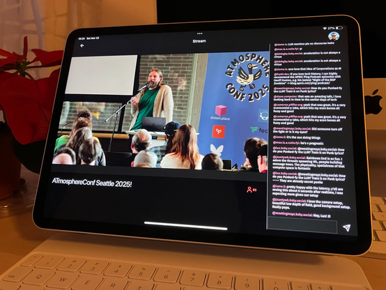 iPad Pro on a keyboard stand on the desk, showing a video from a conference of a man speaking on stage, with a chat sidebar on the right. There's a plant pot and an iMac behind the iPad.