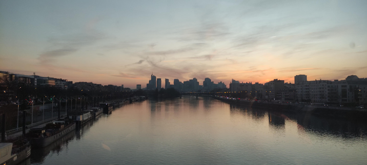 Vue d'un fleuve au crépuscule avec la silhouette des gratte-ciel en arrière-plan. Le ciel se teinte d'orange et de bleu, et les lumières de la ville commencent à briller. Des péniches sont amarrées le long des quais.