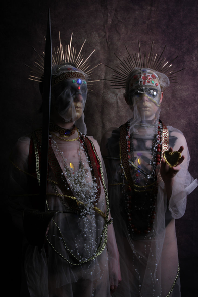 Two figures stand close together in semi-darkness lit by a single light source above them to the right. The figure on the left is at a 45 degree angle to us and holds a sword upright in their right hand, the figure on the right is facing us directly and holds a small heart-shaped mirror in their left hand, directly pointed at us.

Both figures are draped in gauze-like material, wearing headdresses in the forms of sun crown halos, with golden rays forming semi-circles above their heads. Beneath the gauze the top half of their faces/heads are covered with jewel-encrusted masks, and their lower faces seem golden. They have jewels, necklaces, and embroidered ribbons of cloth draped around their necks and covering their chests.