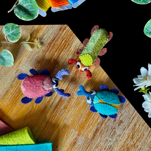 Three different felt beetles, facing each other in a triangle formation, photographed on a black and wooden background with a few decorative plants and pieces of felt.  There is a long green and orange beetle, a round blue and orange one, and an oval pink and blue one (it has a bright green belly)