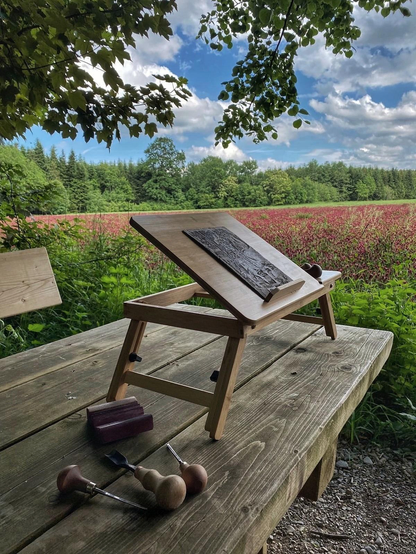 A progress shot of the shown lino block still on the workbench outside with tools strewn to the left of it in the foreground. Behind,  lies a field of flowers backed by a forest.