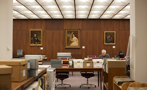 A photo of the archive reading room. There are several square-shaped lights on the ceiling, and a wood-paneled wall at the rear with several large portrait paintings. In the room there are several tables with archival boxes and magazine cases.