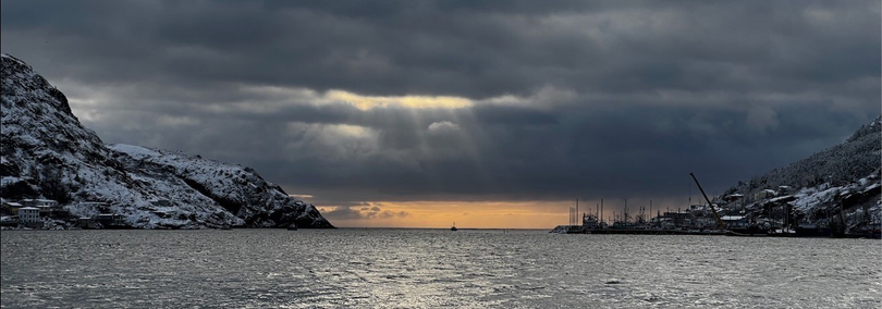 A small fishing boat sails into a cloudy sunrise through The Narrows, St. John's (Canada). The hills are dusted with snow, and rays of sun shine through the dark clouds.