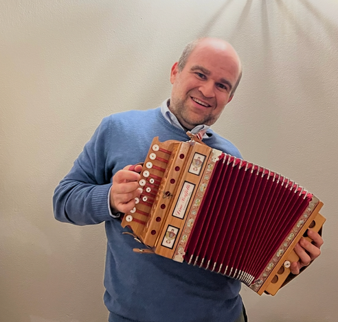 A smiling man in a blue sweater playing a wooden diatonic button accordion with red bellows, standing against a plain light-colored wall.