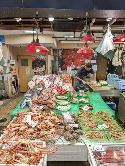 A fish market stall selling a wide variety of fresh fish and seafood.  Prices in Japanese yen are labelled in black ink over white sheets of paper. There is a man cutting and preparing fish behind the table.