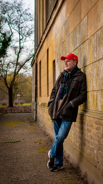 A full-length portrait of an upper-middle-aged man in an orange ballcap, tartan scarf, and winter coat, leaning up against the wall of a crumbling palace in the autumn (this would be me)