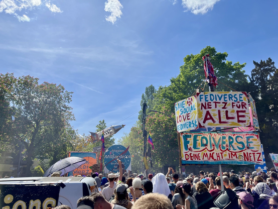 Ein buntes Plakat auf einer Demo, mitten in einer feiernden Menschenmenge: "Fediverse – Netz fuer alle" und "Deine Macht im Netz" steht gross darauf. Andere Schilder fordern "Save Social Love Fediverse" und nehmen ironisch Bezug auf Elon Musk. Zwischen Konfetti, Sonne und Protest zeigt sich hier ein klarer Wunsch: ein offenes, dezentrales Internet jenseits von Algorithmenmacht, Profitinteressen und Plattformkontrolle. Das Fediverse steht dabei als Symbol fuer digitale Selbstbestimmung und Gemeinschaft – getragen von Nutzer:innen, nicht von Konzernen.