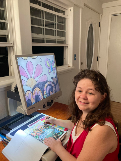 A short white woman sitting in front of a computer-screen-like device with an enlarged drawing of flowers on the screen.  The book, and her hand holding a colored pencil are down on a tray under the monitor and camera.