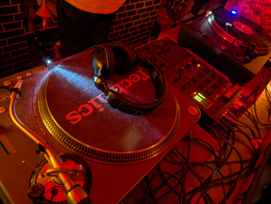 photo of a turntable in the front, visually set in a club setting, with a 3 channel mixer and a second turntable in the background. lighting is cozy reddish. there's a dj headphones on the turntable in the front, but no vinyl.