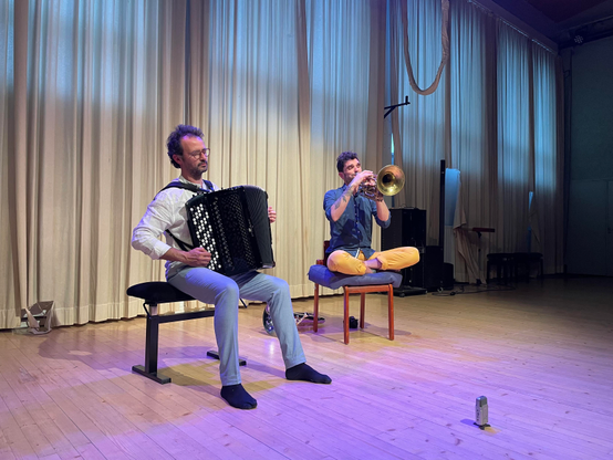 Harmonikansoittaja istuu pianotuolilla, flugabonen soittaja jalat ristissä tuolilla, kumpikin sukkasillaan. Taustalla puulattiaan asti ulottuvat beiget verhot, joiden läpi kevätillan valo kuultaa. 