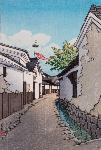 A view looking down a curving lane in an unidentified Japanese town on a bright spring day. On either side of the lane, the plaster walls of the houses looks like they would benefit from some repair. At the other end of the lane, a pair of kids are busy with something unseen; above them, a red carp kite flutters in in the breeze. Overhead, the bright blue sky is populated by a few puffy white clouds.