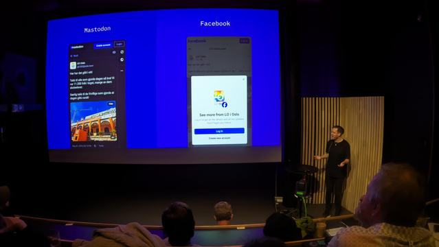 A photo of Matt on stage, pointing towards a screen showing Mastodon and Facebook side-by-side.