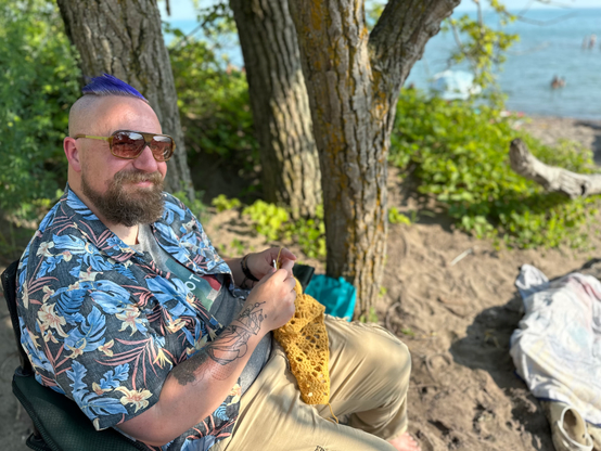 Person sitting on a beach, crocheting. He smiles (It's Will)