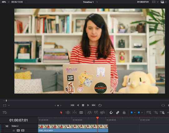 a screenshot from the software DaVinci Resolve showing yours truly (woman with long brown hair wearing a red & white striped shirt) sitting on a couch in front of a computer. The lid is in focus while my face is slightly blurry. There is a bookcase behind me. On the lid of the laptop you can see multiple stickers: "I am part of the rebel alliance" "YunoHost" the Mastodon logo, an illustration of filmmaker Agnès Varda, "We can have a better internet" and "good vibes only". Plushtodon is sitting on the couch next to me