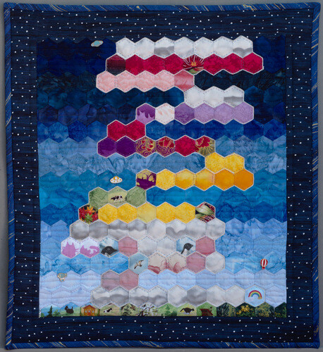 "Morning Mood". Rectangular wall quilt with hexagonal tiles laid out to resemble colourful clouds against a blue sky. Some lapel pins dot the background.