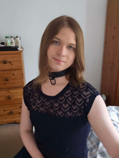 A transfeminine non-binary person sitting on a bed, lit from the right. She's wearing a collar and lace-textured top. 

Her hair is gathered around her shoulder, she's making eye contact with the camera, and she has a slight smile.