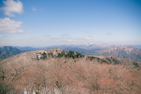 A scenic panoramic view of a mountain range under a mostly clear sky with light clouds. The foreground shows leafless trees with hints of snow, while the background reveals ridges and valleys dusted with snow, stretching into the horizon.