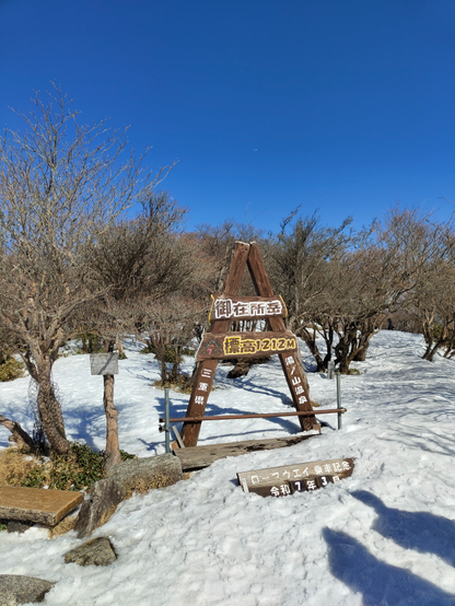 A wooden triangular summit marker stands on a snowy mountain peak under a clear blue sky. The sign reads “御在所岳 標高1212M” indicating Mount Gozaisho at an elevation of 1212 meters, with additional vertical signs showing the location in Mie Prefecture and nearby Yunoyama Onsen. Leafless trees and light snow surround the sign.
