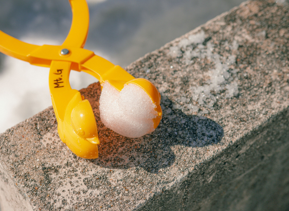 A bright yellow snow mold tool labeled "Mt.G" is resting on a stone ledge. The tool is shaped like a duck and contains a half-formed snow duck, still clinging to the open mold. Scattered snow is visible on the stone surface beneath.
