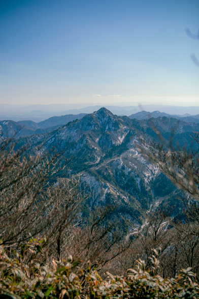 A snow-speckled mountain peak dominates the center of the frame, surrounded by rugged, forested ridges. Sparse branches and foliage partially frame the view, while a clear sky and soft mountain haze stretch into the distance.