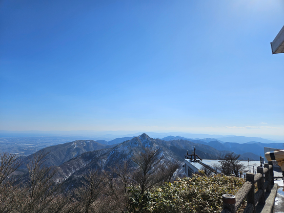 A panoramic view from a mountain lookout shows a snow-dusted ridge extending into a distant mountain range under a clear blue sky. In the foreground, leafless trees and evergreen bushes line a stone-paved area next to a railing and building structure, partially visible on the right.
