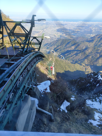 A steep cable car station with a track descends a mountain slope, overlooking a vast cityscape and coastline in the background. The support structure of the cable car is prominent in the foreground, with patches of snow and rocky terrain scattered along the hillside.