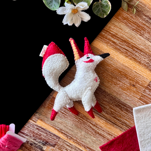 Picture of a handmade felt plushie on a black and wooden background, surrounded with decorative plants, felt and threads. 
The plushie is of an elegant and slender white fox with big red ears. The tip of its tail and feet are red as well and its nose is black. 
Around its eyes, it has elegant red embroidered markings, and 3 softer red dots on its chest. 