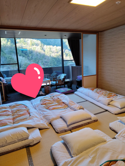 Traditional Japanese tatami room with several neatly arranged futons and pillows on the floor, a large window revealing a forested mountain view, and a pink heart sticker partially censoring the picture.