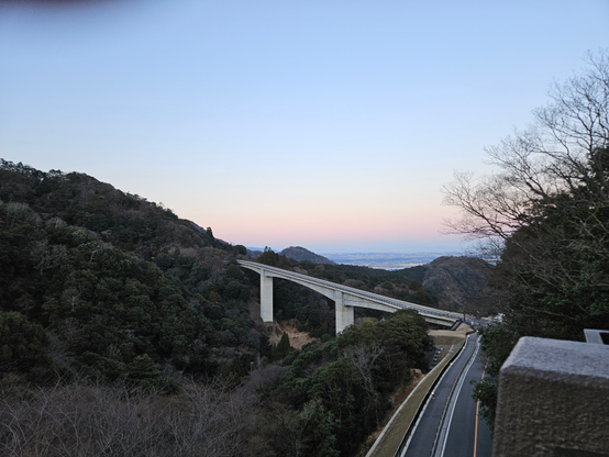 The view from our window. An elevated concrete bridge cutting across a lush, forested mountain valley at sunset, with a winding road below and a gradient sky transitioning from soft orange near the horizon to pale blue above.
