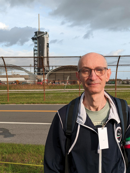 Daylight photo of a bald man with eyeglasses, wearing a green t-shirt and a navy blue sweatshirt, and standing outdoor in front of a metallic mesh fence. Behind the fence is a launch pad with a white rocket and a black service tower next to it.