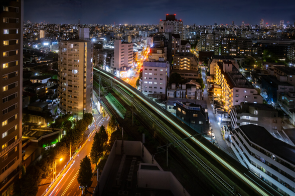 View on Tokyo at night from a skyscrapper building, using a slow shutter speed. A train is crossing the image in diagonal creating a green glow on the black tracks. Buildings and streets around it are bathed in orange or blue lighting depending on the types of lamps used.
