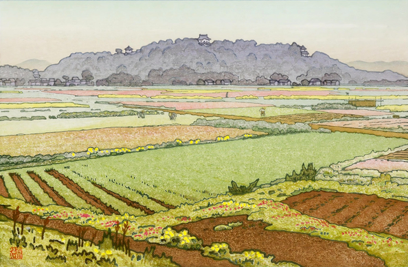 A view across a patchwork of cultivated fields toward Hikone Castle, which stands on a forest-covered hill in the distance. Some of the fields are green with flowers and tidy rows of spring growth, while ither sections are still bare earth. A few figures in broad hats are working in the fields. Beyond, the forest covering the low hill of Mount Konki is dim and shadowed; the highest parts of Hikone Castle reach above the treetops. Overhead, the sky is cloudy and hazy.
