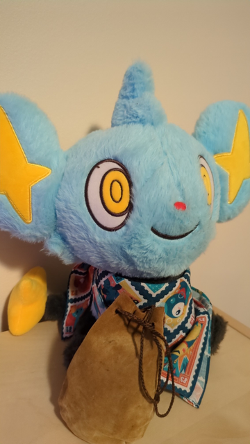 A Shinx plushie wearing a patterned scarf. They have a brown dice bag.