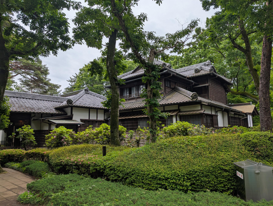 Exterior view of a very fancy Japanese home from the 1930s or earlier. Well trimmed bushes and lush trees partly obscure the house, which has many gables and partly gables roofs made of wavy tiles 