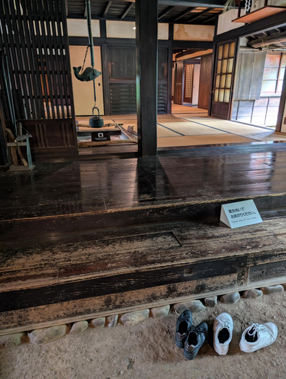 Inside of an old (around 1700s) farmhouse of a village administrator. It shows three different types of floor: in the foreground, stone floor for dirty shoes and messy work; then wood steps and walking area for high traffic zones; then tatami mats around an inset fire pit for sitting and relaxing. The fire pit has an iron kettle and a fish shaped pot hook hanging above it. 