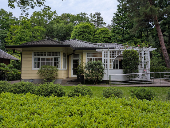 Exterior view of a 1920s Japanese house built with strong European influences. One story, with a simple gabled roof made of tile. The walls are pale yellow slats with white framed windows. A trellis covers a porch on the side 