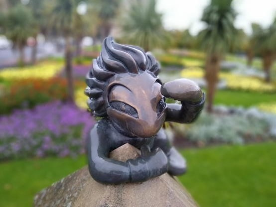 Close up of a bronze statue of a lemming from the computer game "Lemmings". The lemming is clinging with one hand to the top of what seems to be a pillar, and looking past the camera into the distance, shading their eyes with the other hand. Blurred background suggesting a garden landscape with flowers and palm trees.

Lemming statue at Airlie Place in Dundee, artist Alison Conway