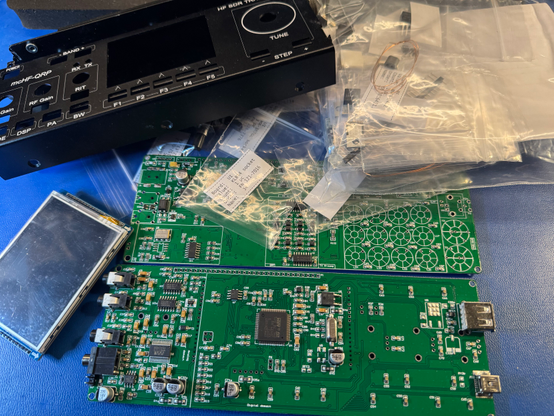 Two green PCB. Lower one is the user interface and the upper one is the RF board. In the right upper corner are little baggies full of parts. In the left upper corner is the enclosure. In the left lower corner is the display for the radio.