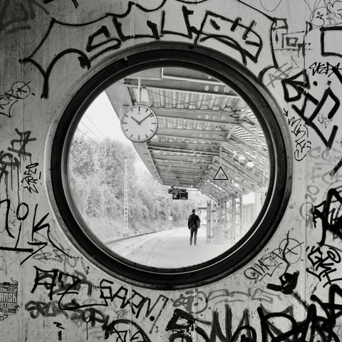 Das Bild zeigt einen Bahnsteig durch ein rundes Fenster in einer mit Graffiti bekritzeln Wand. Die Graffiti sind abstrakt, die Tags kaum zu entziffern. Durch das Fenster sieht man einen Bahnsteig mit einem modernen, metallenen Dach. Eine analoge Bahnhofsuhr ist sichtbar und zeigt 10:09. Im Hintergrund sind Bäume neben dem Bahnsteig zu sehen. Auf dem Bahnsteig, der sich in die Ferne erstreckt, steht eine einzelne Person mit dem Rücken zum Betrachter. Er scheint, den Bahnsteig entlang zu schauen – möglicherweise auf eine Anzeige. Die Person trägt dunkle Kleidung und einen Rucksack. Die Atmosphäre ist still, einsam und etwas melancholisch, mit einem Kontrast zwischen der rauen, urbanen Ästhetik der Graffiti und der ruhigen, fast stillen Szene auf dem Bahnsteig. Das Foto vermittelt ein Gefühl von Isolierung und dem Warten auf etwas.
The picture shows a train platform through a round window in a wall scrawled with graffiti. The graffiti is abstract, the tags barely decipherable. Through the window you can see a platform with a modern, metal roof. An analog station clock is visible and reads 10:09 and in the background you can see trees next to the platform. On the platform, which stretches into the distance, a single person stands with their back to the viewer. The person appears to be looking along the platform - possibly at an information display. The person is wearing dark clothing and carrying a rucksack. The atmosphere is quiet, lonely and somewhat melancholic, with a contrast between the rough, urban aesthetic of the graffiti and the calm, almost silent scene on the platform. The photo conveys a sense of isolation and waiting for something.