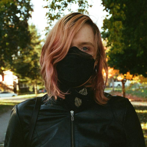 A girl wearing a black leather jacket and a black kn95 mask. She has strawberry blonde hair covering half of her face. In the background is a sunny autumnal park.