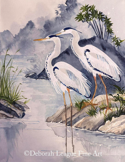 Elegant Watercolor Art Print of two herons at Water's Edge.  Two tropical birds with striking blue and white plumage and long legs stand near the edge of a body of water, surrounded by rocks and sparse vegetation. The background features a misty, wooded landscape, giving a sense of tranquility to the scene.