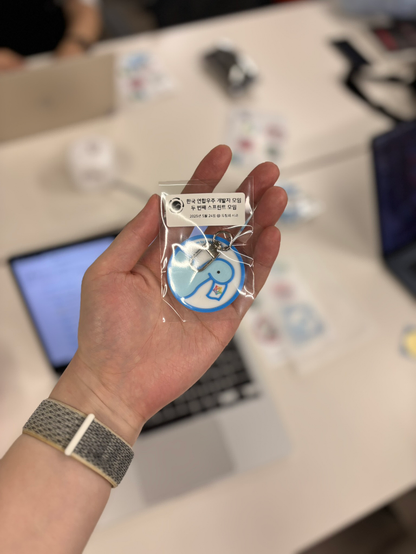 A hand holding a 3D-printed keychain featuring the Fedify dinosaur mascot logo in blue and white colors. The keychain is packaged in a clear plastic bag with Korean text indicating it's from the FediDev KR Second Sprint on May 24, 2025. The background shows laptops and stickers on a table, suggesting a coding workspace at the sprint event.