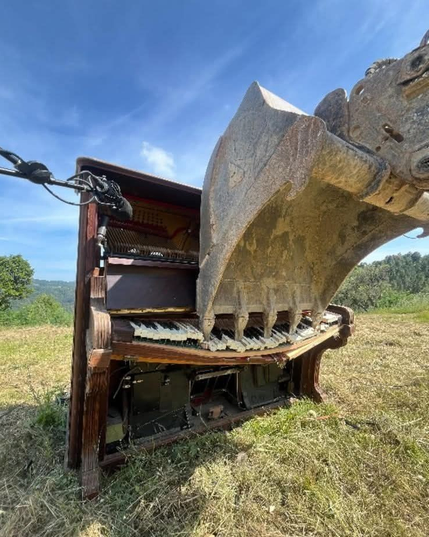 bulldozer bucket playing a piano that it is actually destroying, in a field