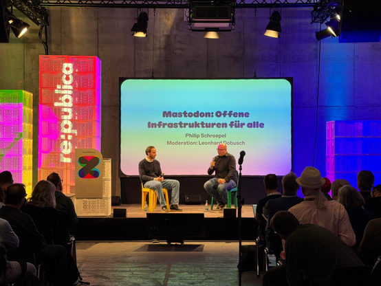 A stage at re:publica - two men sitting in front of a white slide with the words "Mastodon: Offene Infrastrukturen fur alle".