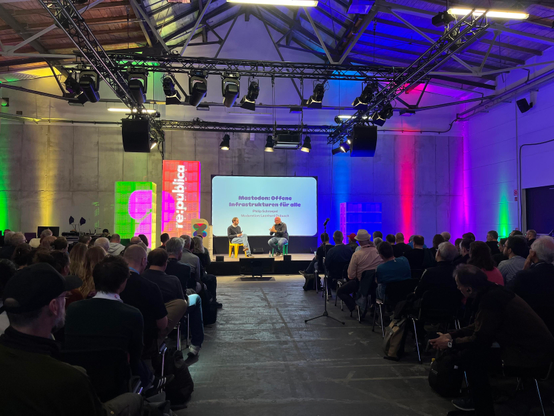 A long shot of a stage at re:publica - in the distance, two men sitting in front of a white slide with the words "Mastodon: Offene Infrastrukturen fur alle". The photo is taken along the central aisle, with rows of attendees on either side.