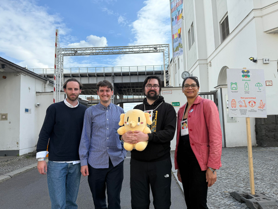 Members of the Mastodon team, 3 men and a woman, holding a beige plush Mastodon mascot, near some industrial looking buildings. The sky is blue with white fluffy clouds.