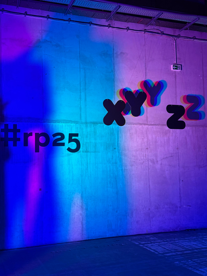The re:publica hashtag rp25 on a concrete wall lit with neon blue and pink lights, with the letters X, Y, and Z hanging and casting shadows nearby.